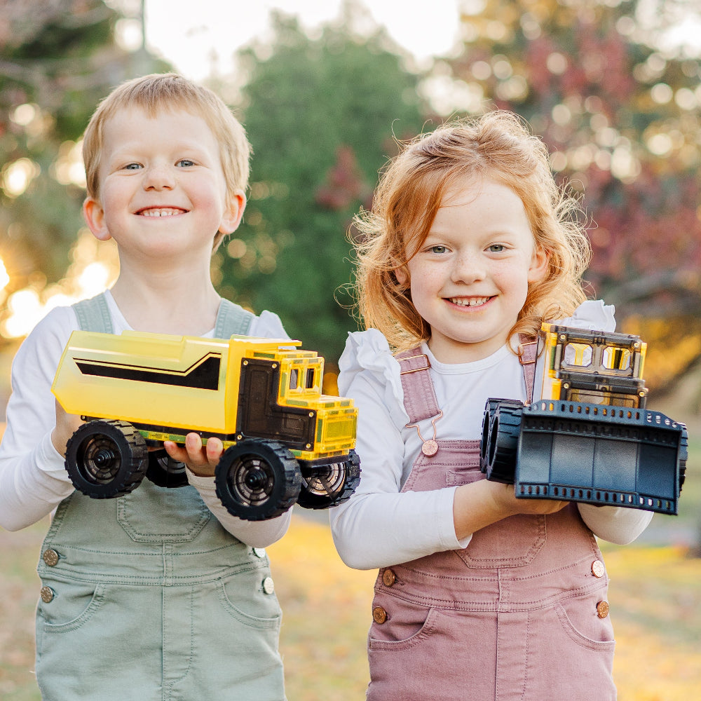 Two children holding magnetic tile toy trucks outdoors with a blurred natural background