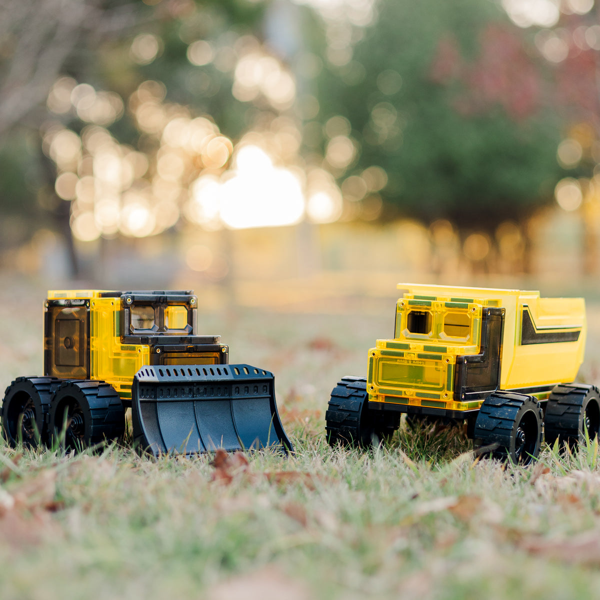 Two magnetic tile toy construction vehicles on grass with a blurred natural background