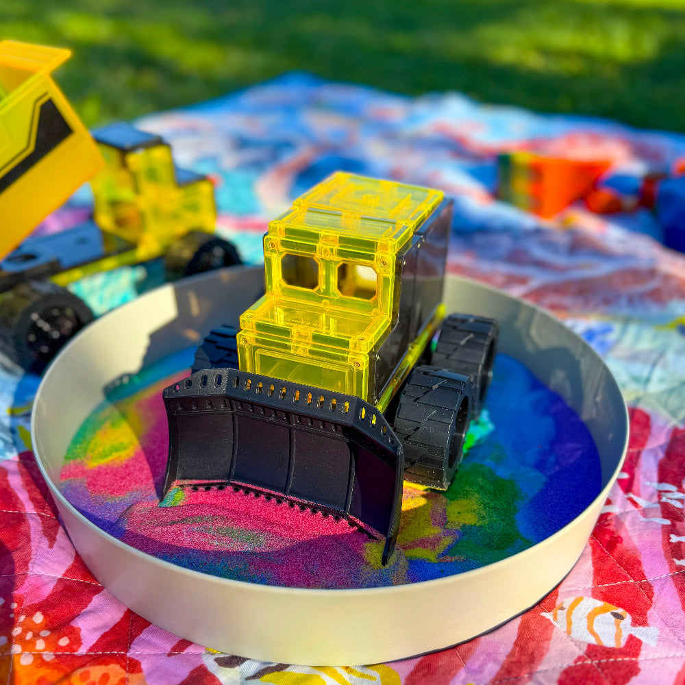 Magnetic tile bulldozer on a tray with rainbow sand with a grassy background outdoors